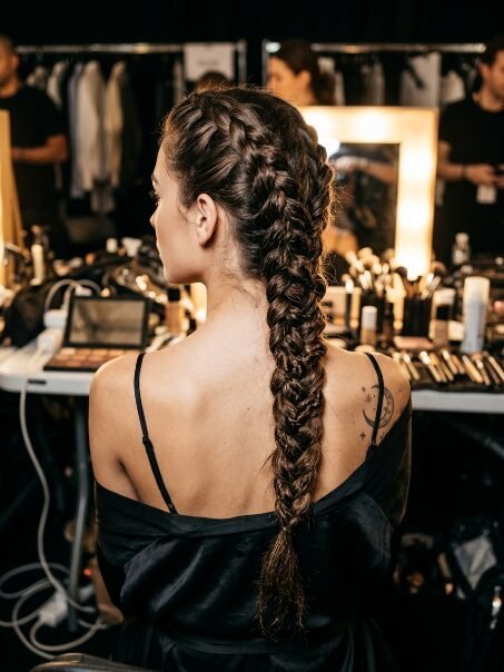 Back view of a woman with long brown hair styled in a single thick Dutch braid, seen in a makeup studio.