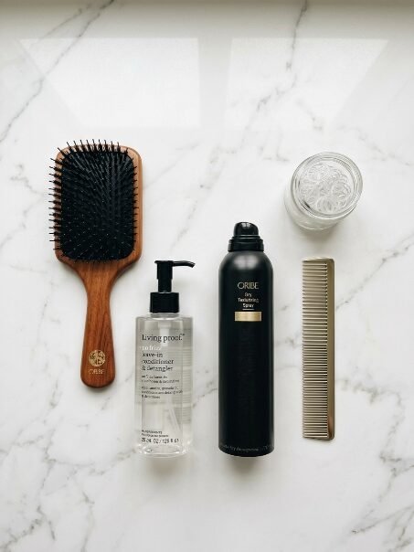 Wooden hair brush, Oribe spray, Living Proof conditioner, and hair ties on white marble.