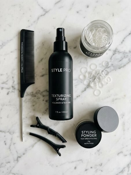 Flat lay of hair styling spray, powder, comb, clips, and clear elastic bands on a white marble surface.