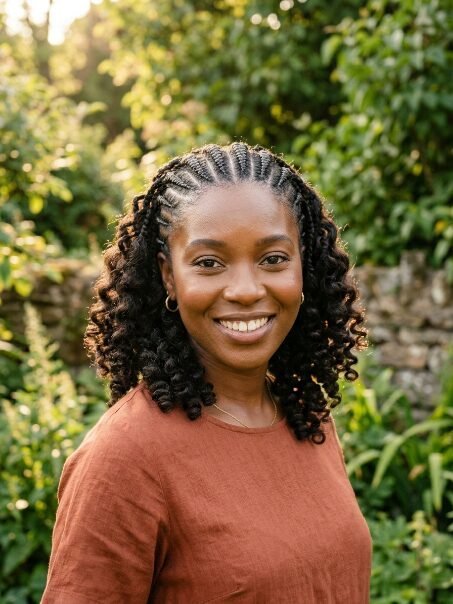 Portrait of a smiling Black woman with cornrows and curly hair wearing a terracotta top outdoors in sunlight.