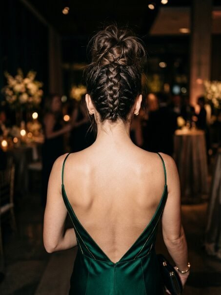 Back view of a woman with a braided bun hairstyle wearing a backless emerald green formal dress.