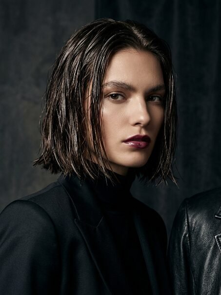 Close up of a woman wearing a black blazer and dark lip color with a wet hairstyle against a dark background.