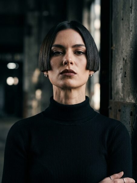 Portrait of a woman with a dark bob haircut and ribbed black sweater looking intensely at the camera.