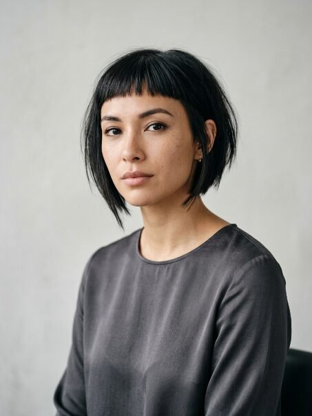 Close-up portrait of a woman with dark hair styled in a short bob against a neutral grey wall.