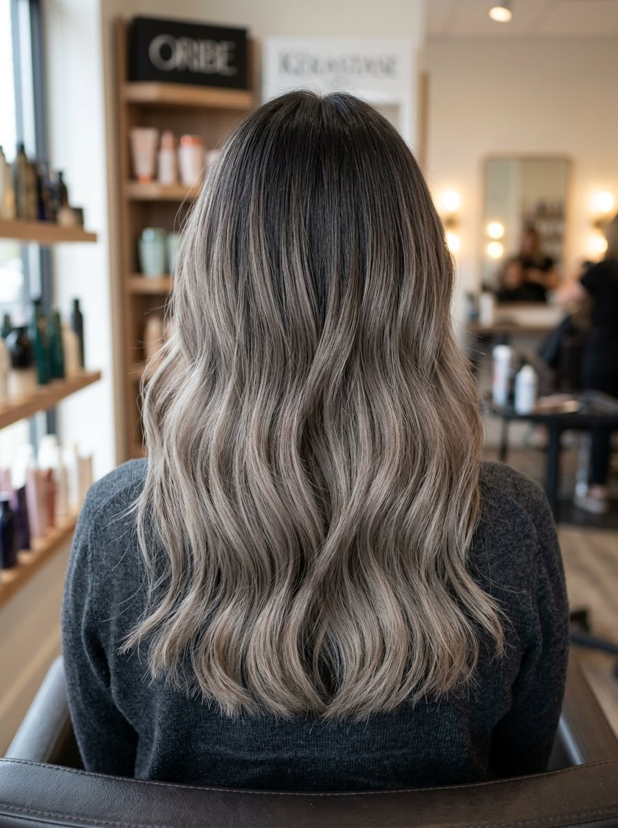 Long wavy hair with dark roots and ash blonde balayage styled in a salon chair.