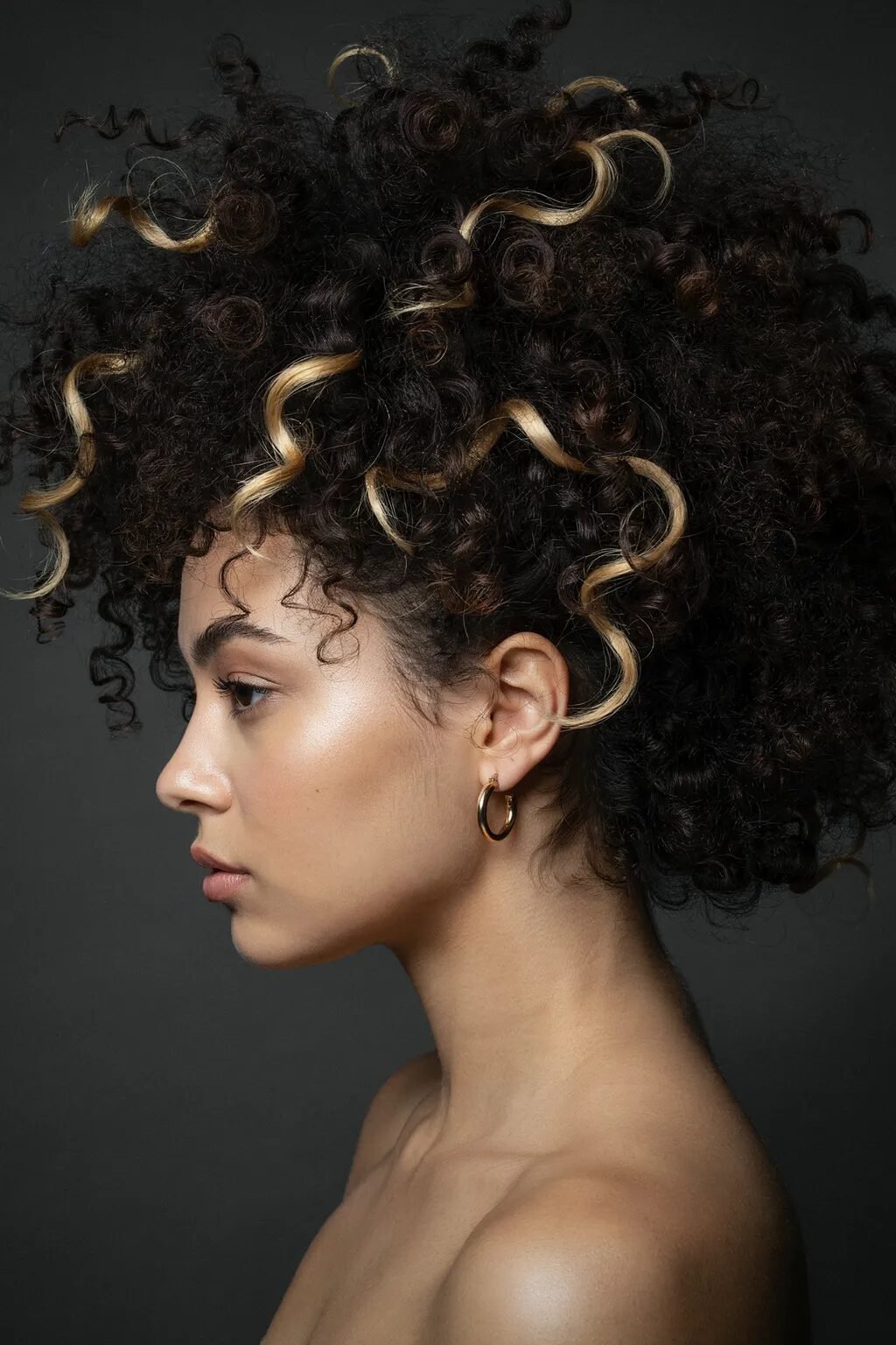 Side view of a woman with dark curly hair and blonde highlights against a neutral grey background.