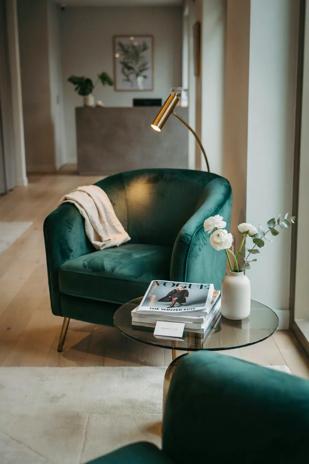 A plush green velvet tub chair with a throw blanket next to a glass table holding magazines and a vase of white flowers.