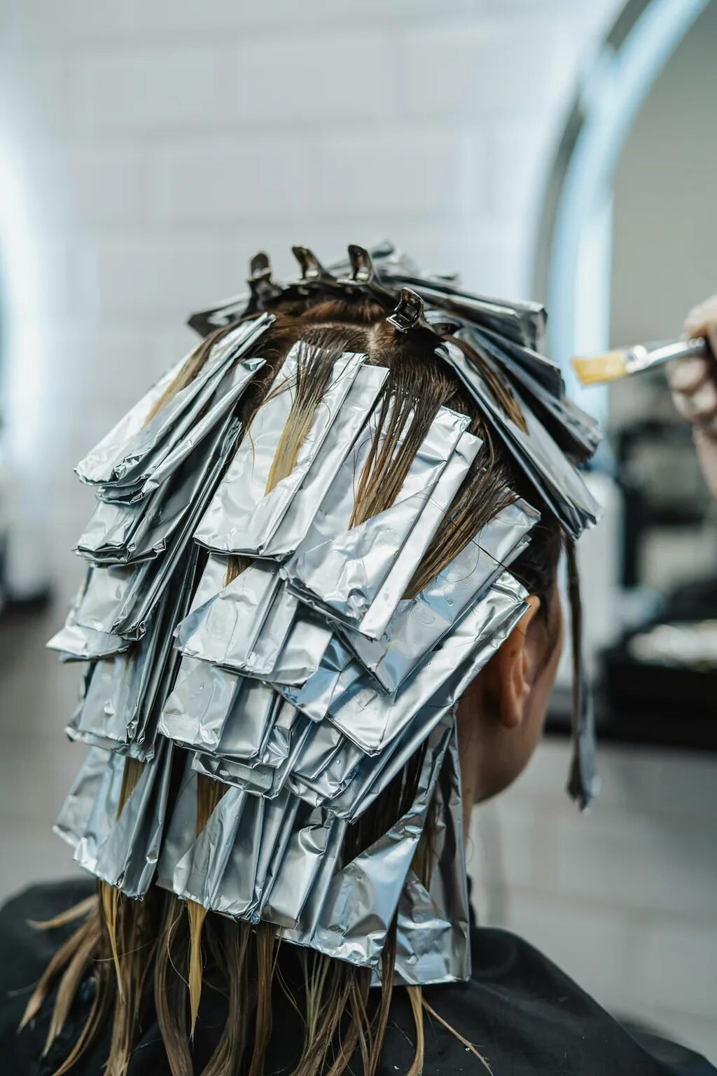 Close-up back view of a woman with sections of hair wrapped in silver foils at a hair salon.