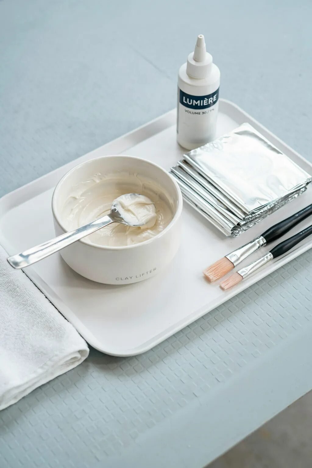 Professional hair dye bowl with cream lightener, foil sheets, developer bottle, and application brushes on a tray.