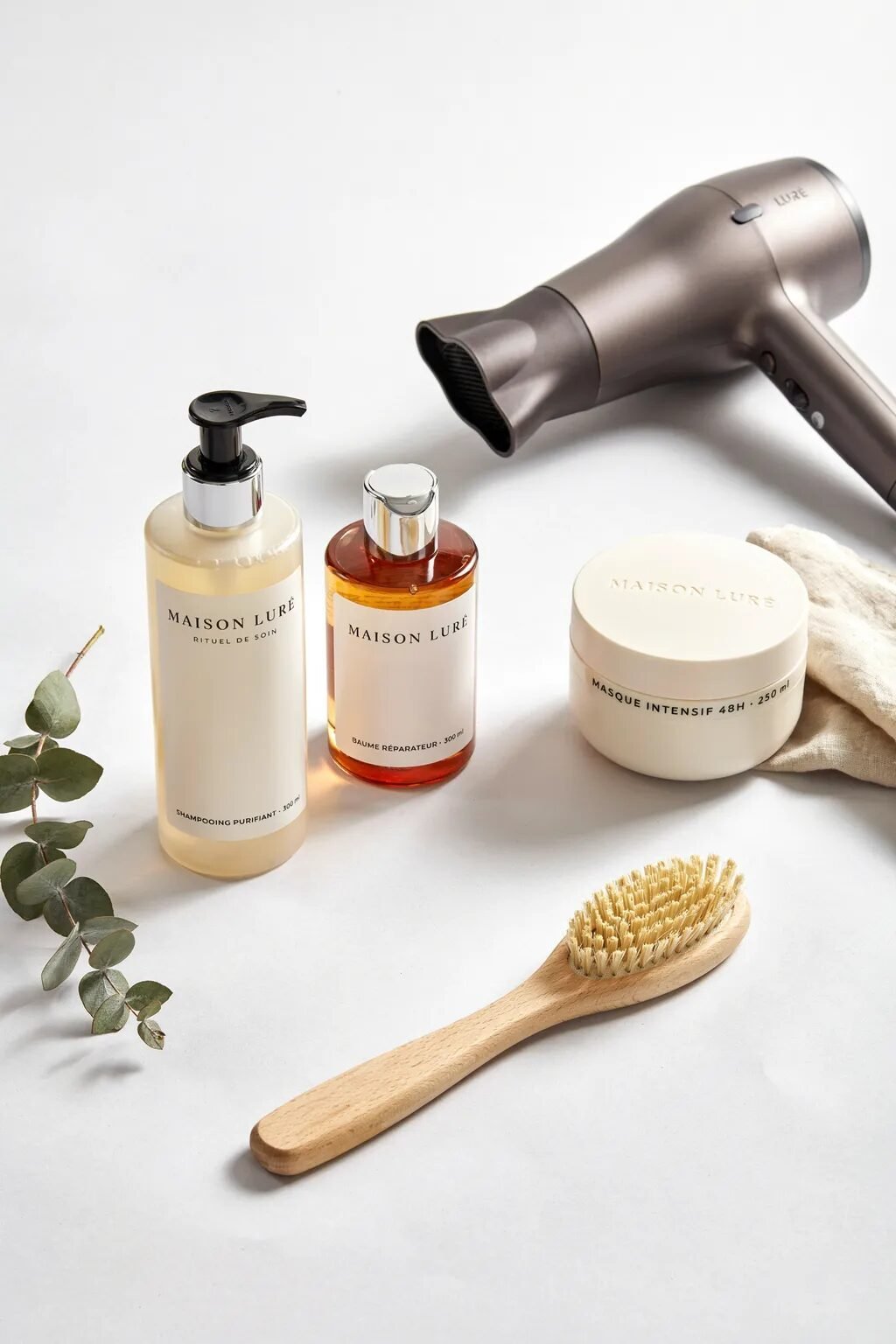 Collection of Maison Lure shampoo, conditioner, mask, hair dryer, and wooden brush on white background.