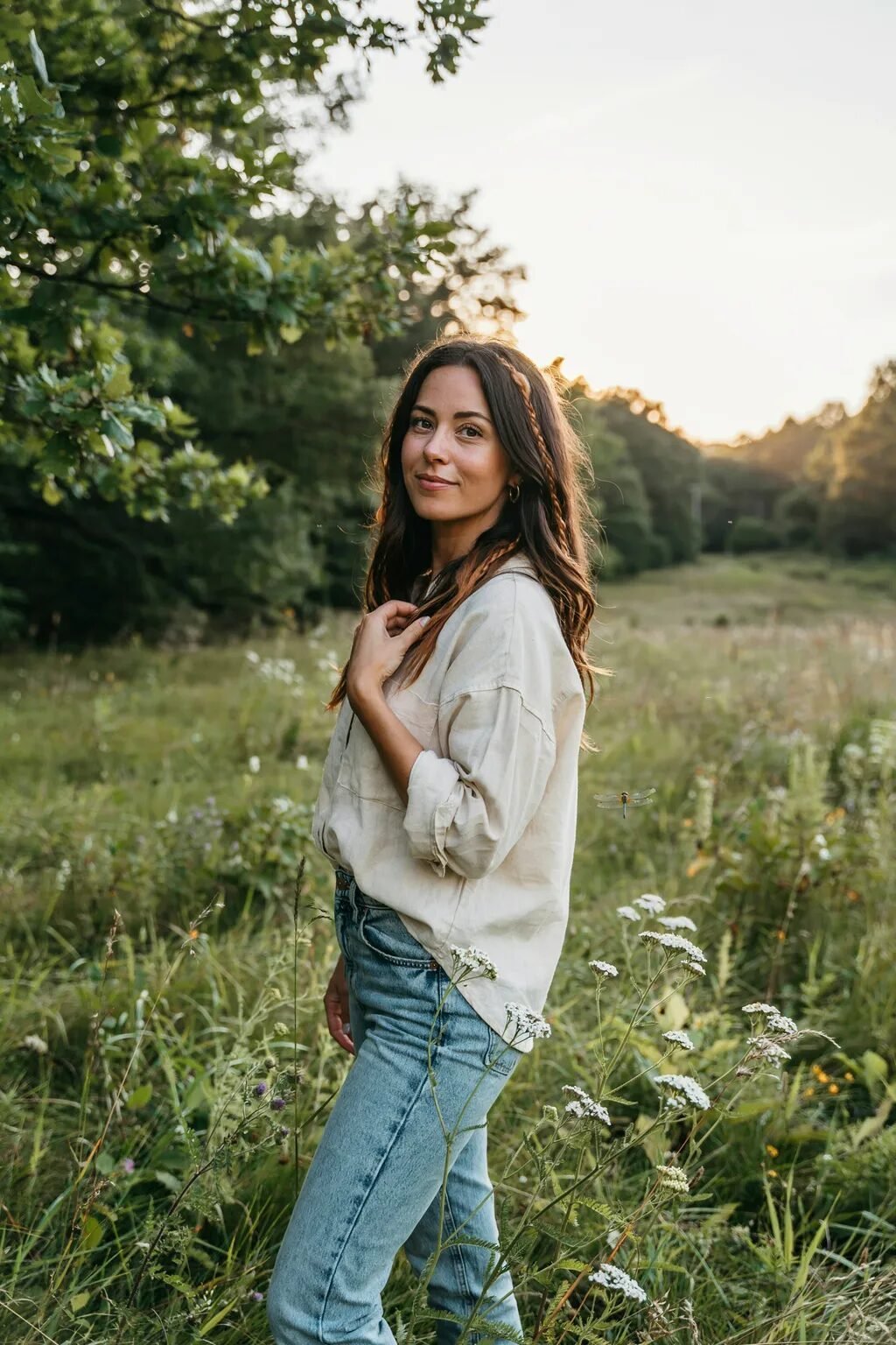 A smiling woman with long brown hair stands in a grassy meadow with white wildflowers at sunset.