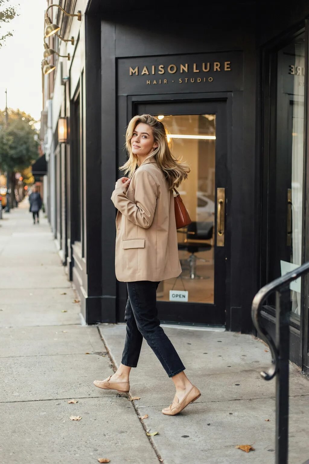 Blonde woman in business casual attire smiling while walking on a city sidewalk in front of a black storefront.